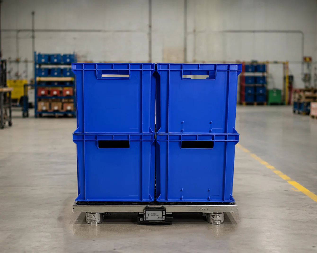Blue plastic storage bins stacked on a smart pallet system in a warehouse with shelving and yellow safety posts on the floor.