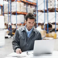 Man working at a laptop in a smart warehouse, with pallets and shelves in the background
