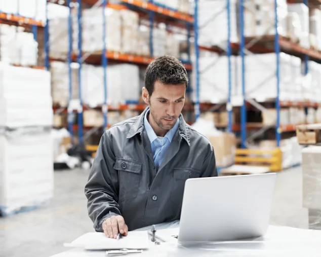 Man working at a laptop in a smart warehouse, with pallets and shelves in the background