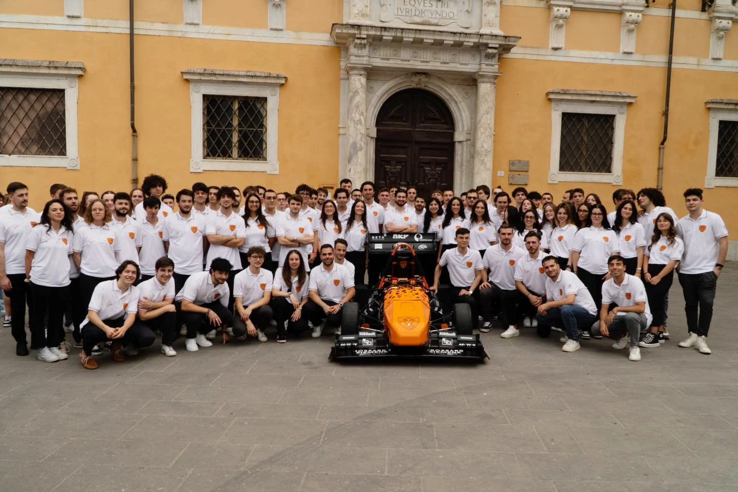 Un grande gruppo in camicie bianche posa intorno a un'auto da corsa arancione davanti a un edificio storico con facciata gialla.