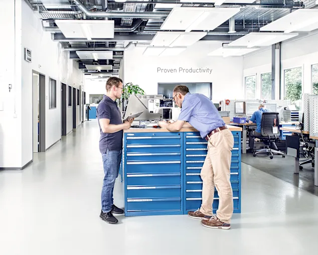 Two men in a test lab examine a sewing machine on a desk in an industrial lab with additional workstations in the background. Key visual for Expert Test Services.