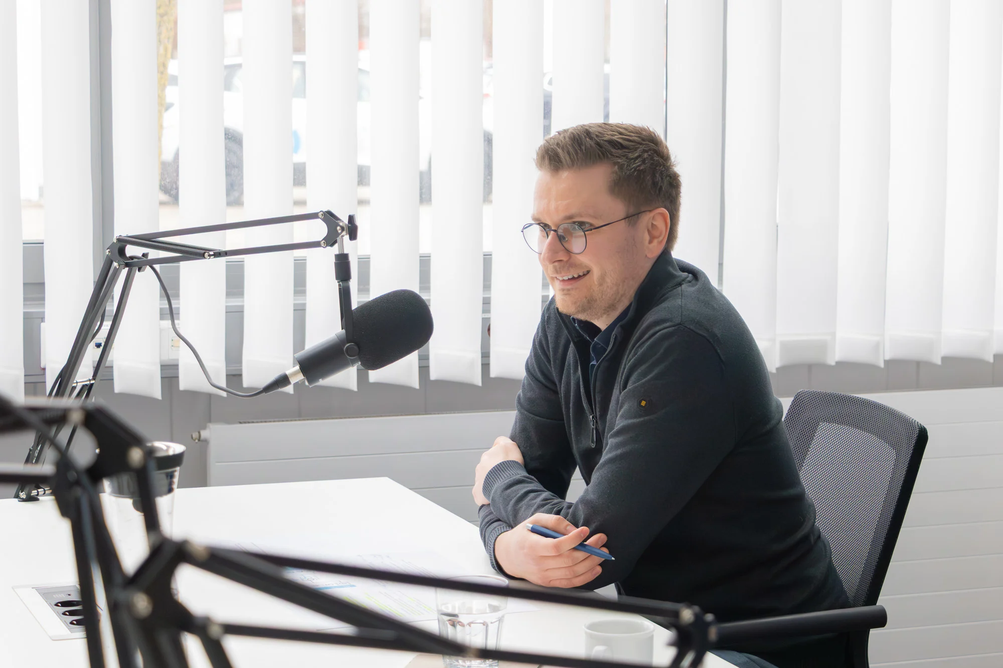 Benjamin Hartinger sitzt in einem hellen Aufnahmeraum und spricht in ein Broadcast Mikrofon auf einem Stativarm.
