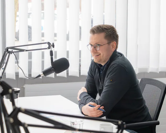 Benjamin Hartinger sitzt in einem hellen Aufnahmeraum und spricht in ein Broadcast Mikrofon auf einem Stativarm.