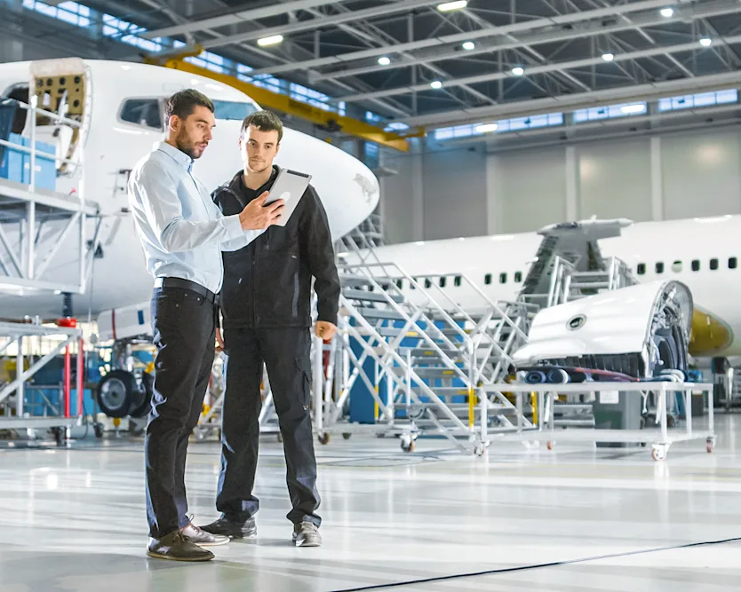I en flyfabrik fokuserer to mænd på en tablet og analyserer information omgivet af flykomponenter.