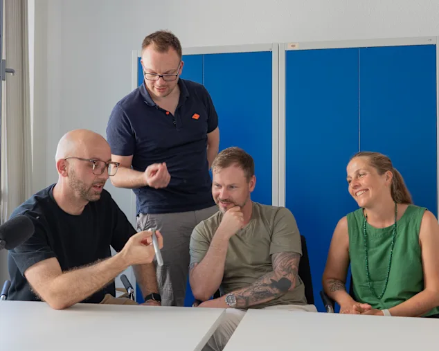 Vier Personen im Gespräch während einer Podcast-Aufnahme zum Thema DIN- und Normteile. Ein Teilnehmer hält ein Verbindungselement in der Hand, während ein Mikrofon im Vordergrund steht.