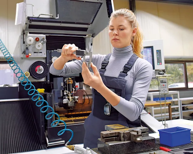Mladá žena pracuje jako technik počítačového numerického řízení. Je vidět na svém pracovišti v modrém overalu, jak měří vnější průměr trubky.