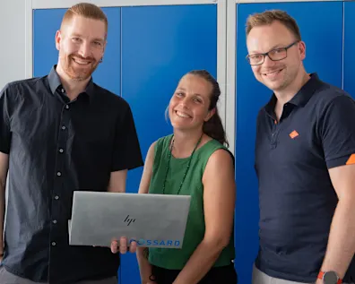 Matthias Schorer, Maja Matzke und Tobias Engelberg stehen lächelnd vor blauen Schränken; Maja Matzke hält einen geöffneten Bossard Laptop.