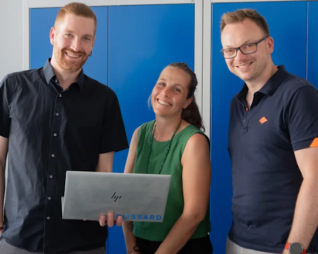 Matthias Schorer, Maja Matzke und Tobias Engelberg stehen lächelnd vor blauen Schränken; Maja Matzke hält einen geöffneten Bossard Laptop.