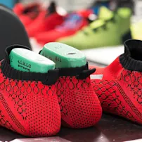 Red and black knitted shoe uppers with green molds inside, placed on a table. Background shows blurred colorful shoes.