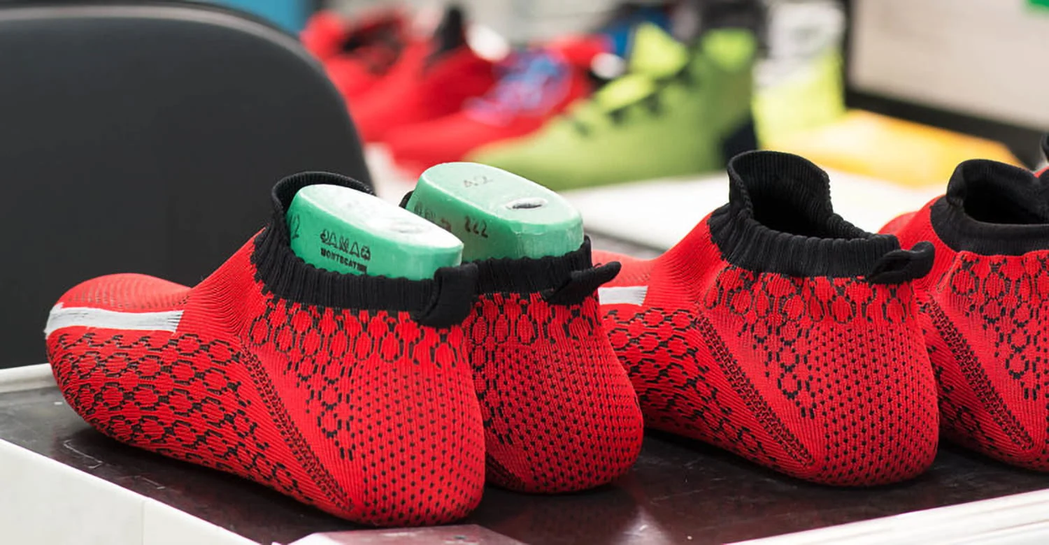 Tomaie di scarpe rosse e nere lavorate a maglia con stampi verdi all'interno, posizionate su un tavolo. Sullo sfondo si vedono scarpe colorate sfocate.