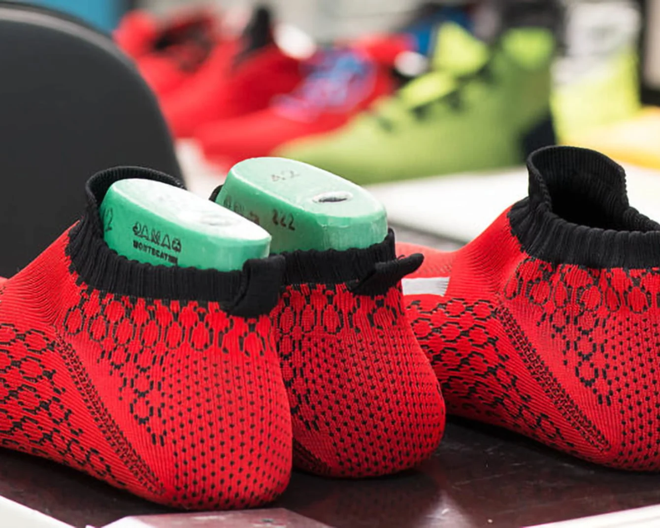 Tomaie di scarpe rosse e nere lavorate a maglia con stampi verdi all'interno, posizionate su un tavolo. Sullo sfondo si vedono scarpe colorate sfocate.