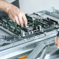 Bàn tay của kỹ thuật viên đang lắp đặt linh kiện trên bo mạch chủ máy tính màu xanh được bao quanh bởi các bộ phận phần cứng và quạt làm mát trên bàn làm việc