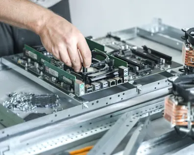 Mano de técnico instalando componente en placa base verde de ordenador rodeada de piezas de hardware y ventiladores de refrigeración en banco de trabajo
