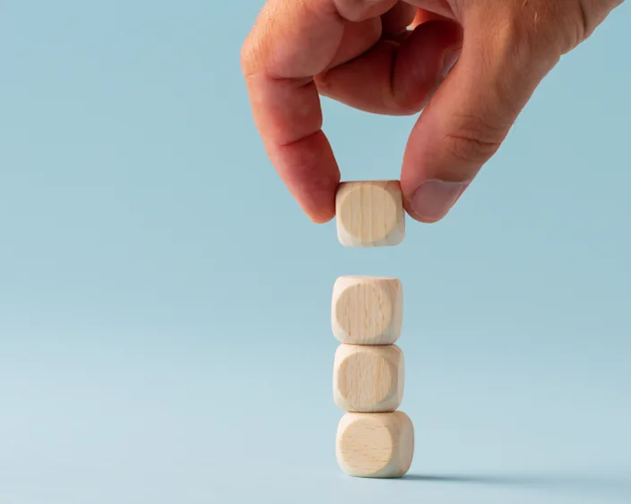 Hand stapelt vier hölzerne Würfel aufeinander vor hellblauem Hintergrund, symbolisiert Stabilität und Wachstum.