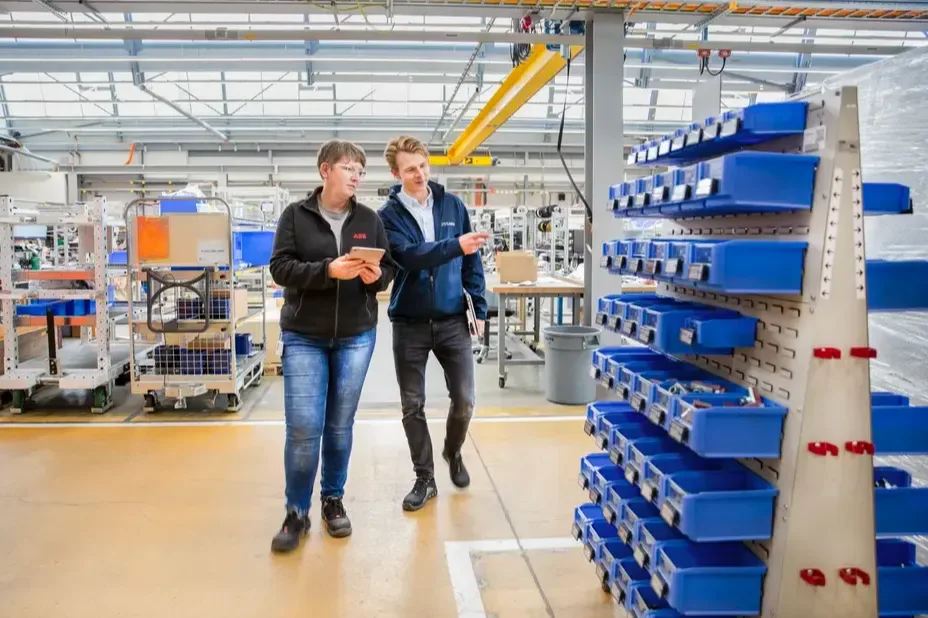 Two people walk through the shop floor, the woman holding a tablet in her hand, the man pointing to a mobile trolley equipped with SmartLabel Cloud bins.