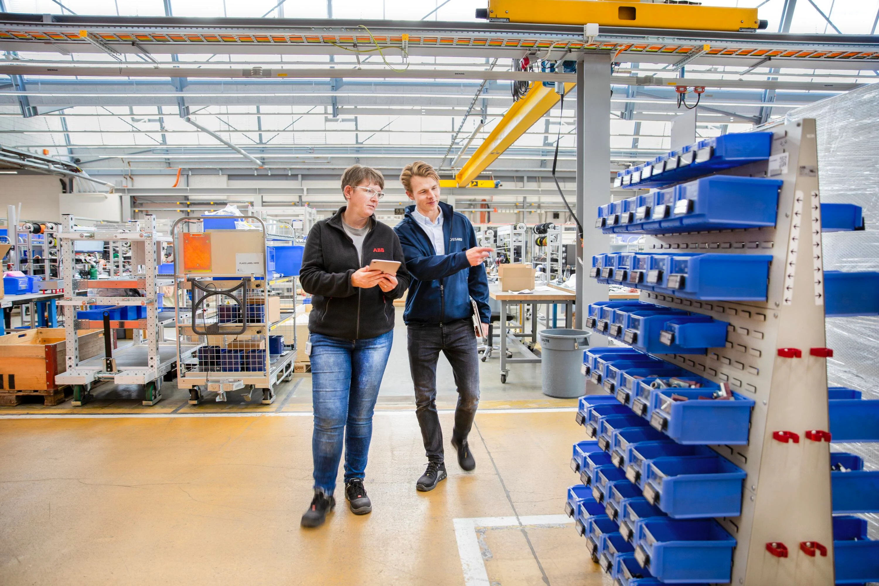 Two people walk through the shop floor, the woman holding a tablet in her hand, the man pointing to a mobile trolley equipped with SmartLabel Cloud bins.