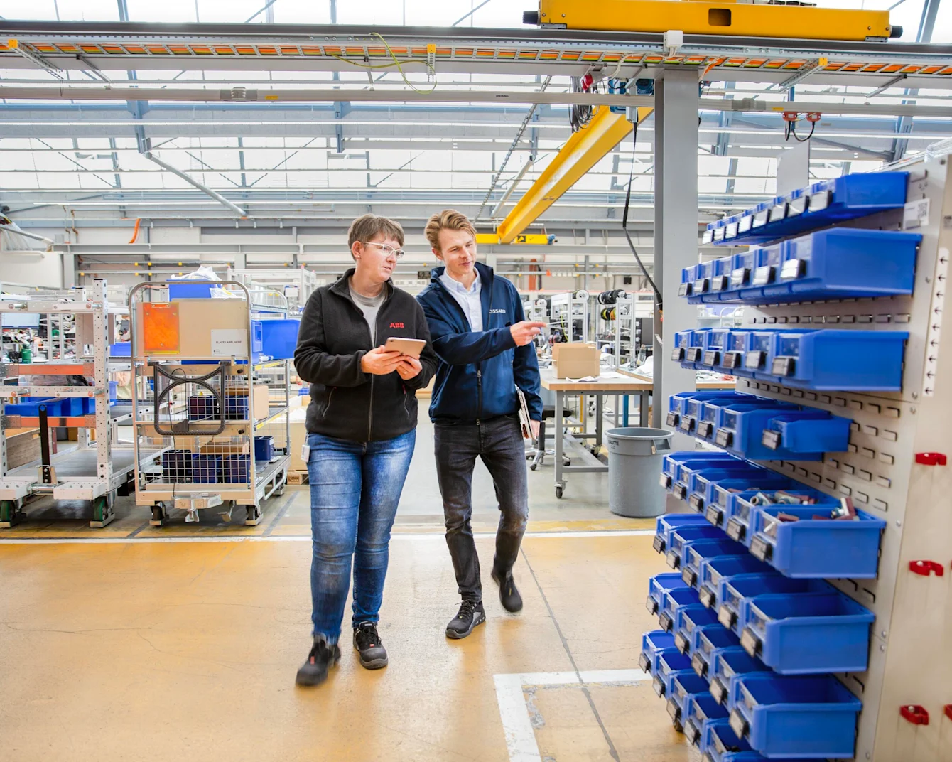 Two people walk through the shop floor, the woman holding a tablet in her hand, the man pointing to a mobile trolley equipped with SmartLabel Cloud bins.