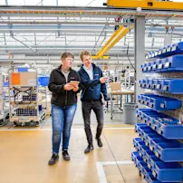 Dwie osoby przechodzą przez halę produkcyjną, kobieta trzyma tablet w ręku, mężczyzna wskazuje na mobilny wózek wyposażony w pojemniki SmartLabel Cloud.
