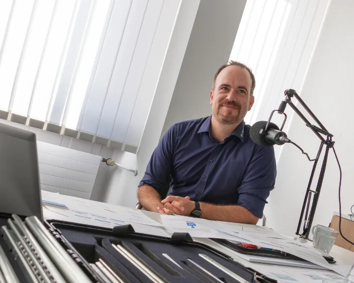 Alexander Dobler mit Mikrofon am Podcast-Tisch, umgeben von Teleskopschienen und Unterlagen, lächelnd vor dem Mikrofon.