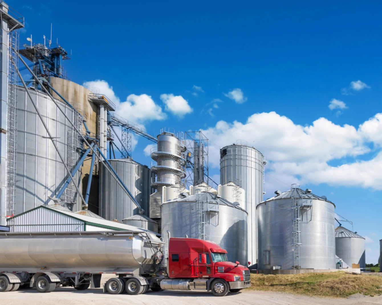 Red semi truck parked in front of large grain silos with open blue sky in the background