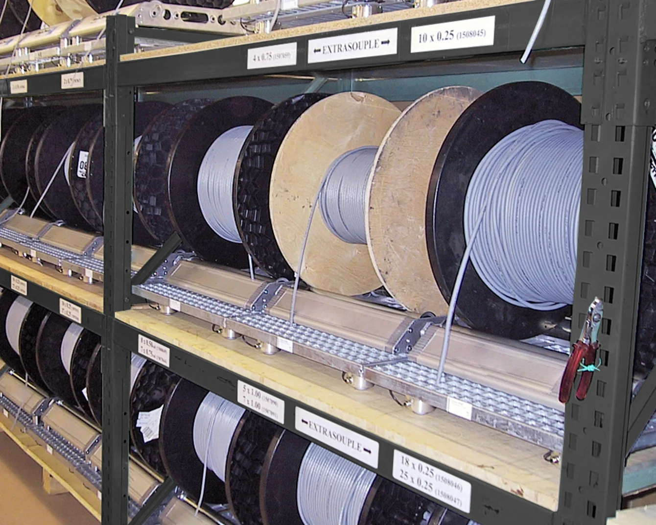 Scaffalatura industriale che contiene grandi bobine di cavi con rocchetti di filo bianco e nero organizzati su scaffali metallici con sezioni etichettate.