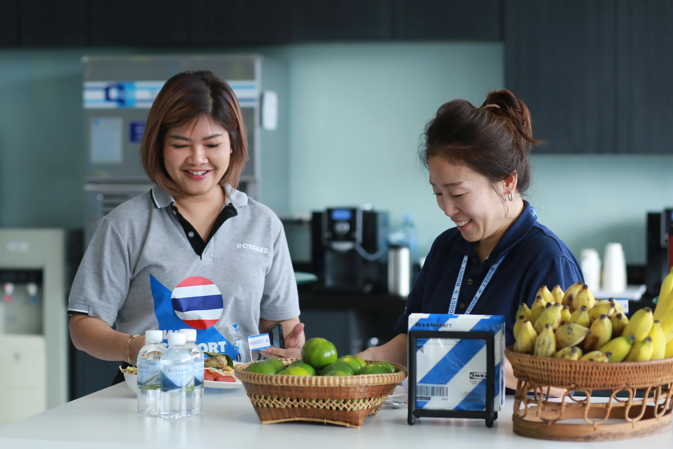 Two women smile joyfully while standing beside a table adorned with an assortment of fresh fruit.