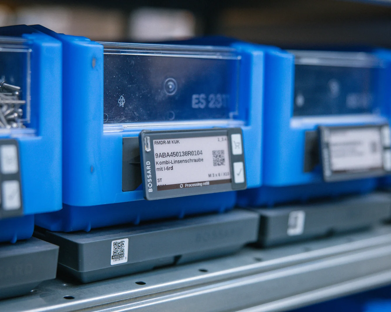 Blue plastic storage bins with clear fronts and digital labels displaying part numbers, arranged on a shelf system.