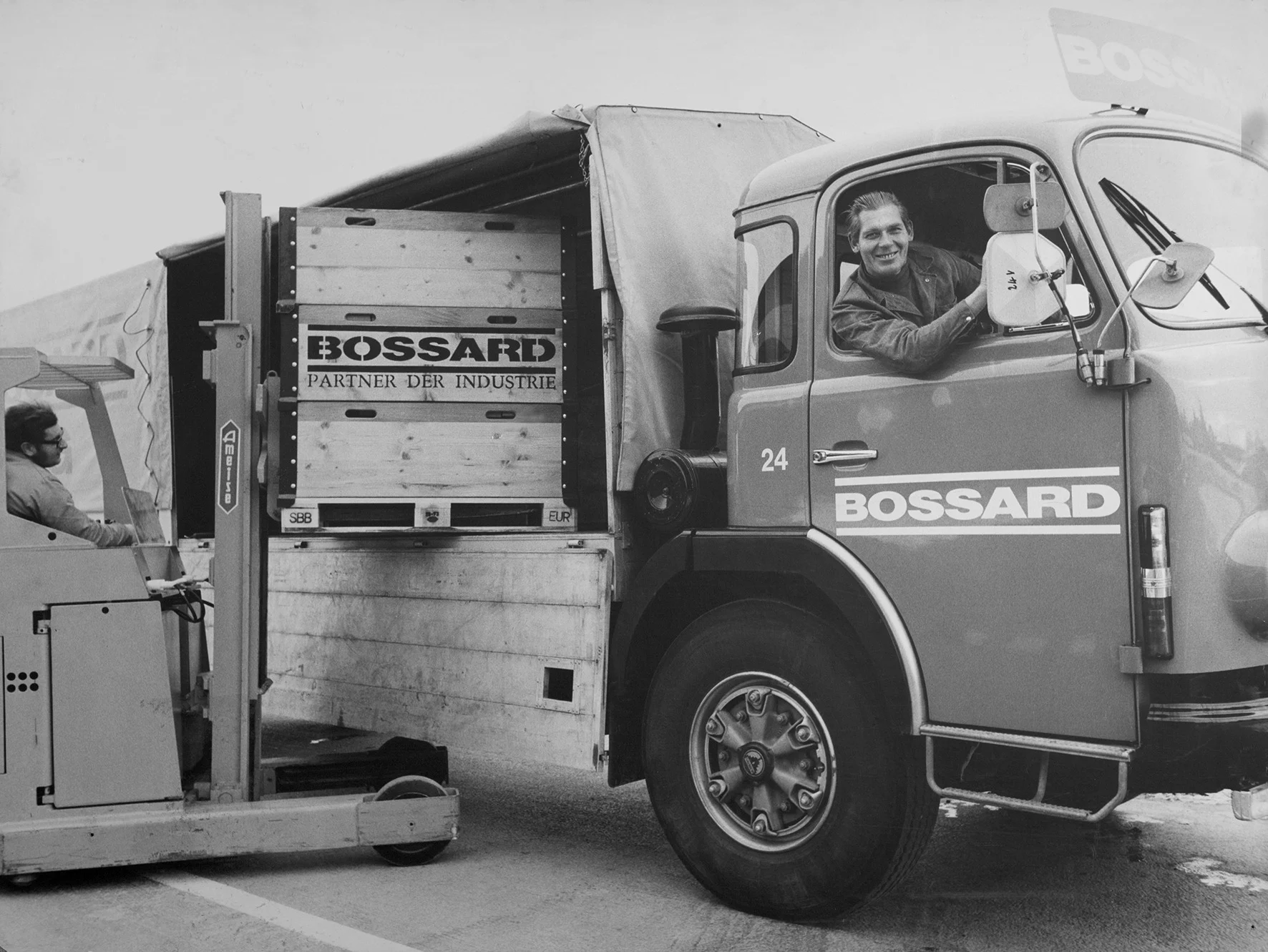 Sort-hvidt billede fra fortiden viser en mand i en stablervogn, der aflæsser en Bossard-lastbil, mens en anden mand inde i lastbilen smiler, hvilket skaber en samarbejdende scene.
