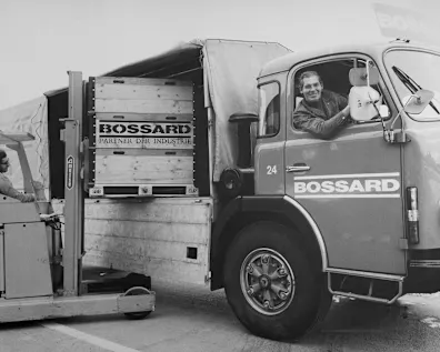 Black and white image from the past showing a man in a stacker unloading a Bossard truck while another man inside the truck smiles, creating a collaborative scene.