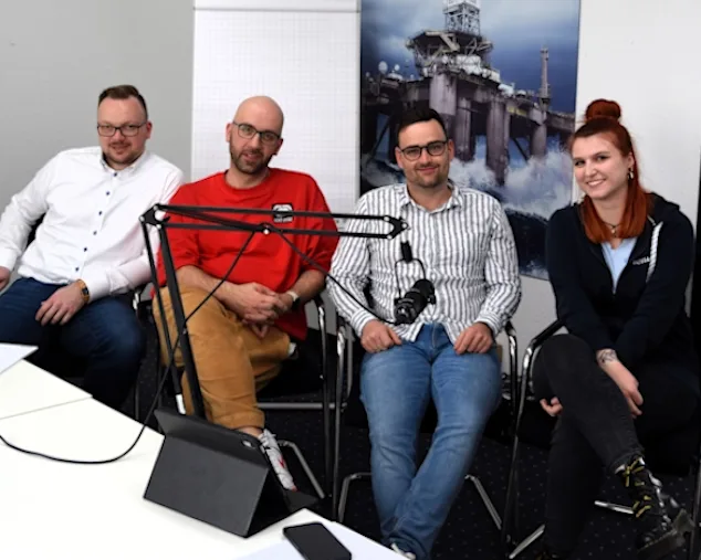 Zwei Podcast-Moderatoren und 2 Gäste sitzen an einem Tisch mit Mikrofonen, im Hintergrund eine Offshore-Plattform