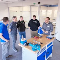 A group of men stand around a workbench with tools and discuss together.