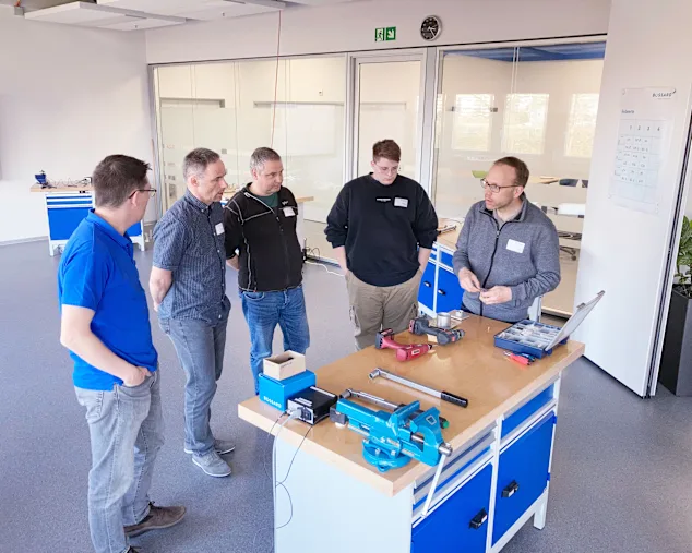 Eine Gruppe von Männern steht um einen Werkbank mit Werkzeugen und diskutieren gemeinsam.