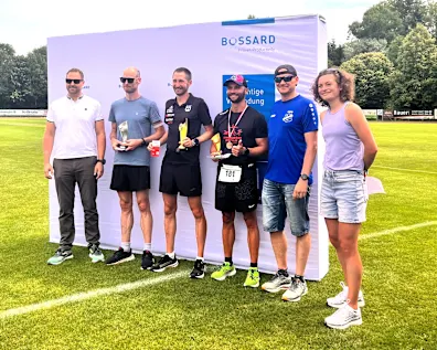 Gruppenfoto von sechs Personen inklusive Lauf-Siegern mit Pokalen vor einer Bossard-Wand auf einem Sportplatz.