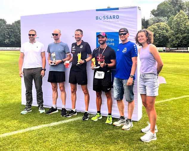Gruppenfoto von sechs Personen inklusive Lauf-Siegern mit Pokalen vor einer Bossard-Wand auf einem Sportplatz.