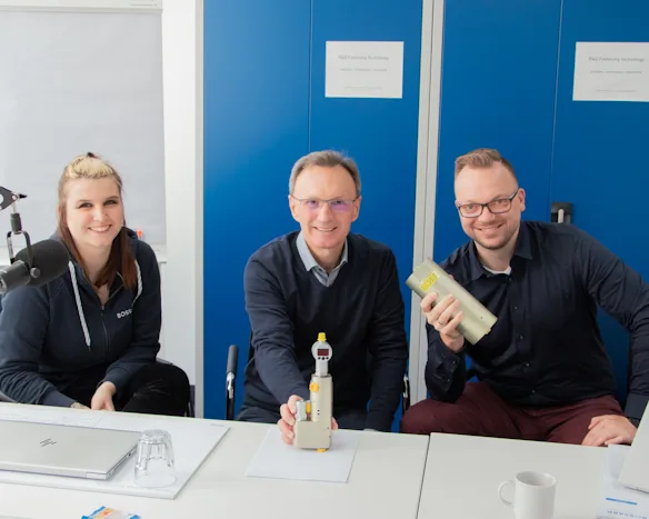 Drei Personen in einer Diskussion, halten MiniBooster-Komponenten, mit blauen Schränken im Hintergrund.