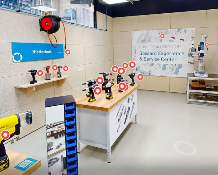 Innenaufnahme des Bossard Experience & Service Center mit verschiedenen Nietsetzgeräten und Werkzeugen auf Werkbänken, Beschilderung „Niettechnik“, interaktiven Markierungen und einem Willkommensbanner an der Wand.