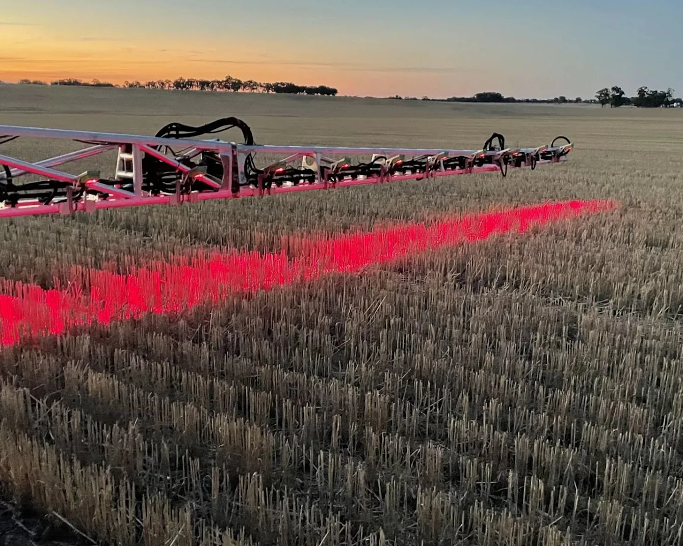 Rode landbouwspuitapparatuur die helder rood licht werpt over een geoogst stoppelveld bij zonsondergang met bomen aan de horizon.