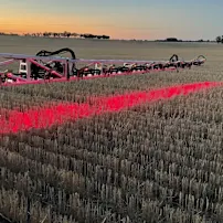 Rode landbouwspuitapparatuur die helder rood licht werpt over een geoogst stoppelveld bij zonsondergang met bomen aan de horizon.