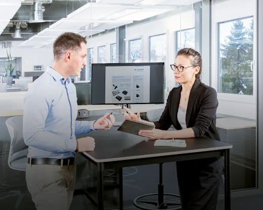Two people at a desk in a modern office discuss fastening a composite material; one holds a sandwich panel, the other a fastener.