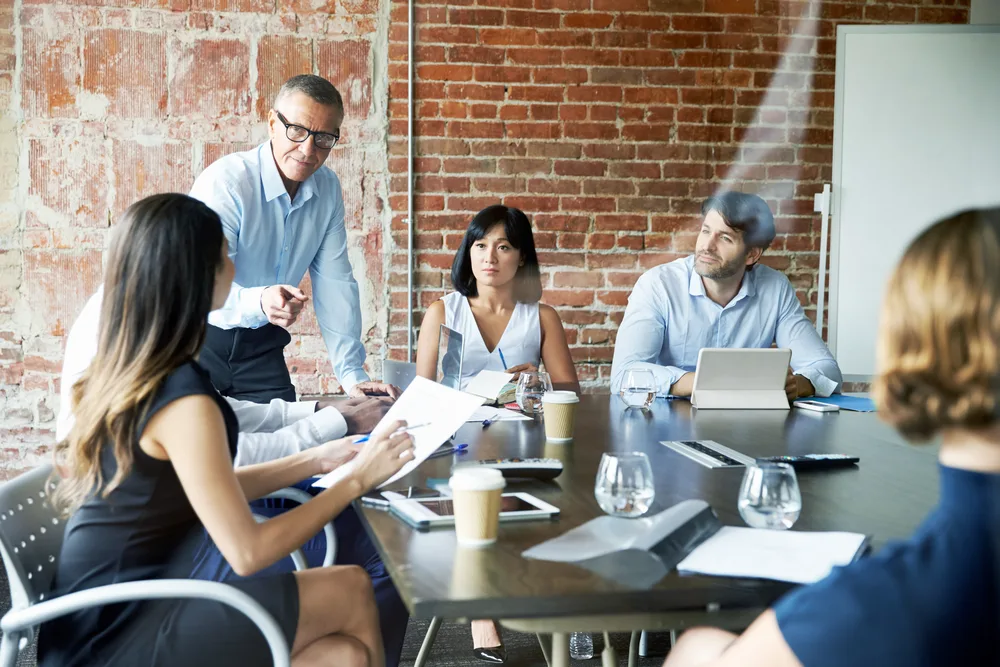 Business situation showing 5 people in a fancy meeting room in industrial design. A senior manager is pointing to a woman with a sheet of paper in her hands, discussing things.
