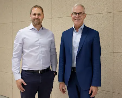 Florian Heuter im weißen Businesshemd auf der linken Seite, Frank Hilgers im blauen Anzug auf der rechten Seite, stehend vor einem hellbraunen Hintergrund.