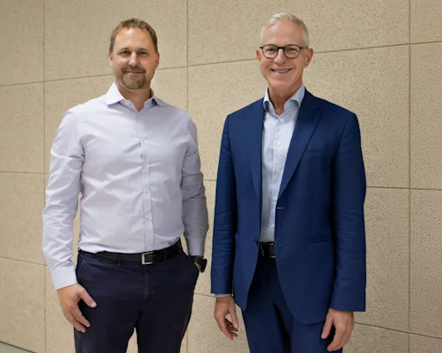 Florian Heuter im weißen Businesshemd auf der linken Seite, Frank Hilgers im blauen Anzug auf der rechten Seite, stehend vor einem hellbraunen Hintergrund.