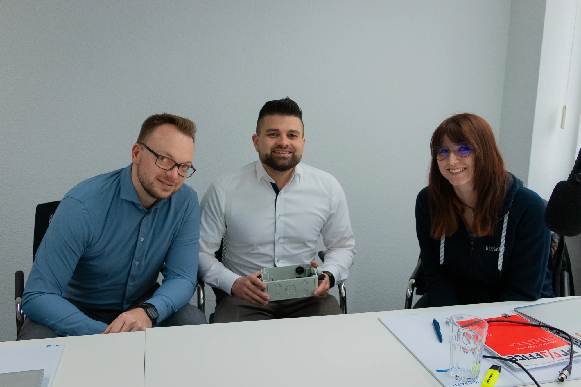 Drei lächelnde Experten sitzen nebeneinander im Podcast Studio, Jan Oelmaier in der Mitte hält eine graue Musterbox mit Powerelementen.