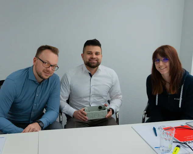 Drei lächelnde Experten sitzen nebeneinander im Podcast Studio, Jan Oelmaier in der Mitte hält eine graue Musterbox mit Powerelementen.