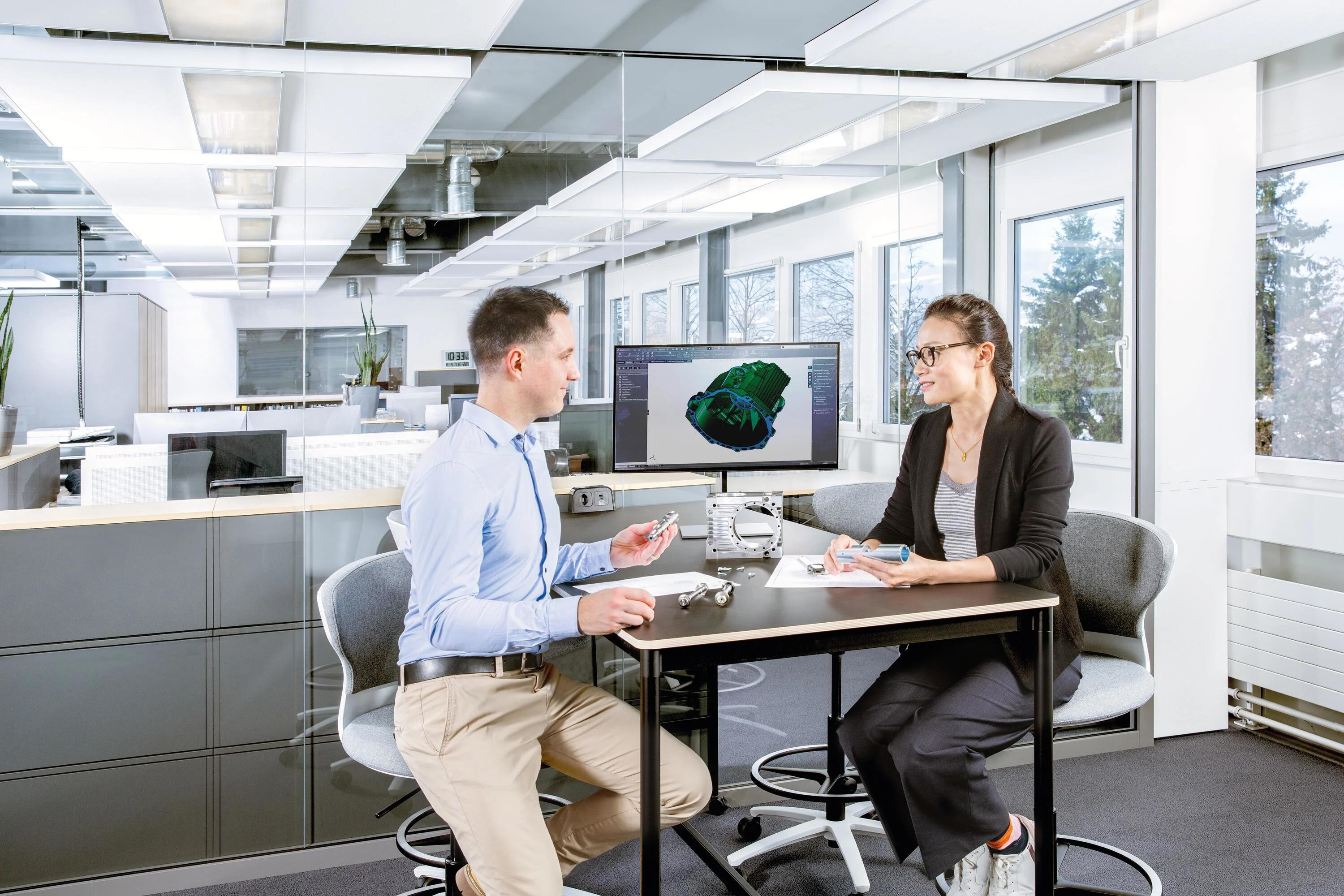 A male and female sit at a table with a screen displaying a technical drawing. On the table are a physical application, a technical drawing, and several screws. Key visual for Expert Design only.