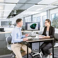 A male and female sit at a table with a screen displaying a technical drawing. On the table are a physical application, a technical drawing, and several screws. Key visual for Expert Design only.
