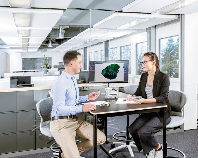 A male and female sit at a table with a screen displaying a technical drawing. On the table are a physical application, a technical drawing, and several screws. Key visual for Expert Design only.