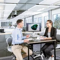 En mand og en kvinde sidder ved et bord med en skærm, der viser en teknisk tegning. På bordet er en fysisk applikation, en teknisk tegning og flere skruer. Nøglevisuel kun for Expert Design.