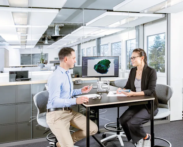 En mand og en kvinde sidder ved et bord med en skærm, der viser en teknisk tegning. På bordet er en fysisk applikation, en teknisk tegning og flere skruer. Nøglevisuel kun for Expert Design.