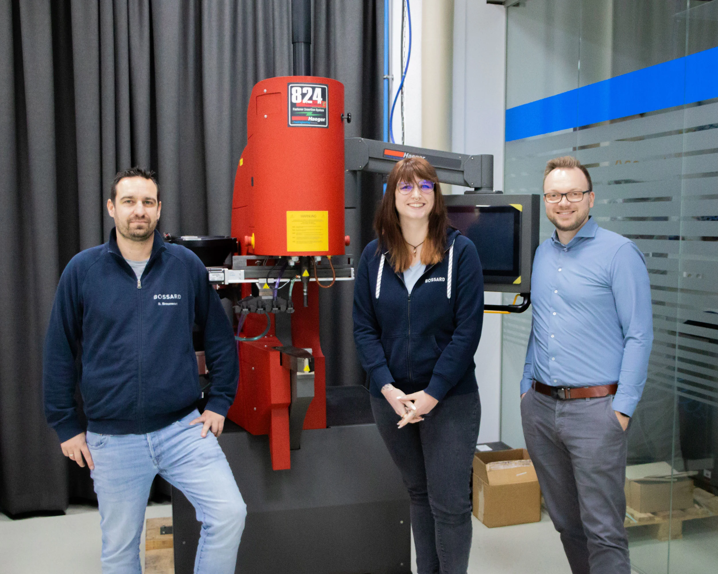 Drei Personen stehen vor einer großen roten Einpressmaschine (Haeger 824) in einem modernen Raum. Zwei tragen Bossard-Kleidung. Alle lächeln in die Kamera. Im Hintergrund sind Vorhänge und Glaswände sichtbar.